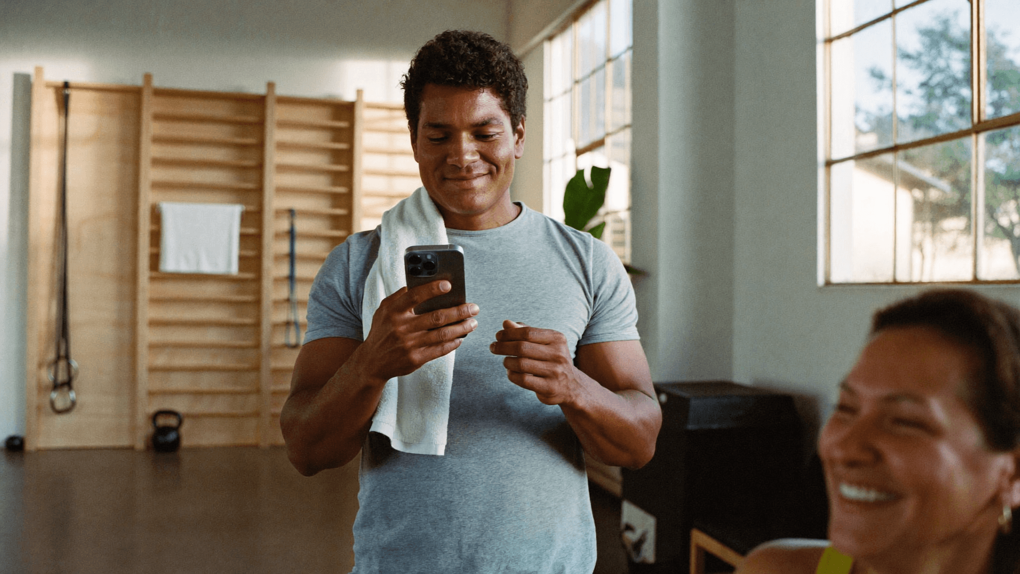 Personal trainer no estúdio com a toalha no ombro, sorrindo ao olhar o celular entre as séries