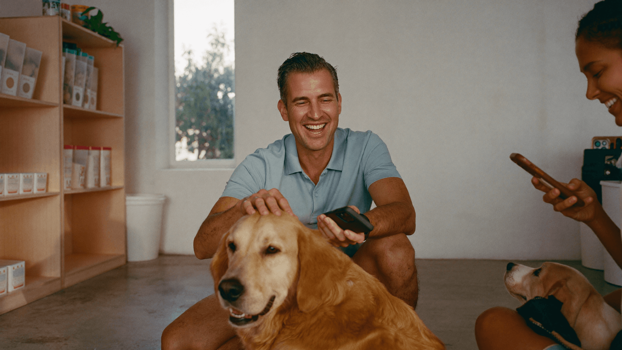 Dono de pet shop agachado fazendo carinho em um golden retriever, com o celular na mão
